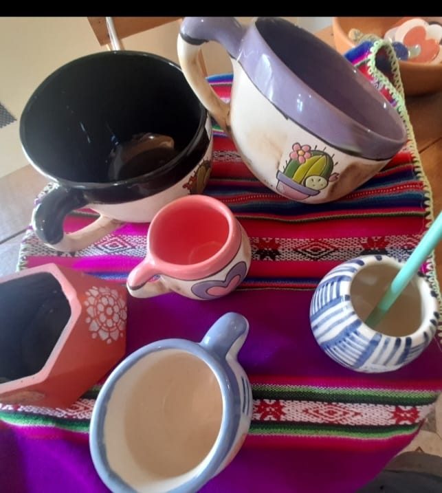 Tazas, una jarrita y un cuenco de cerámica pintados, sobre un aguayo de colores