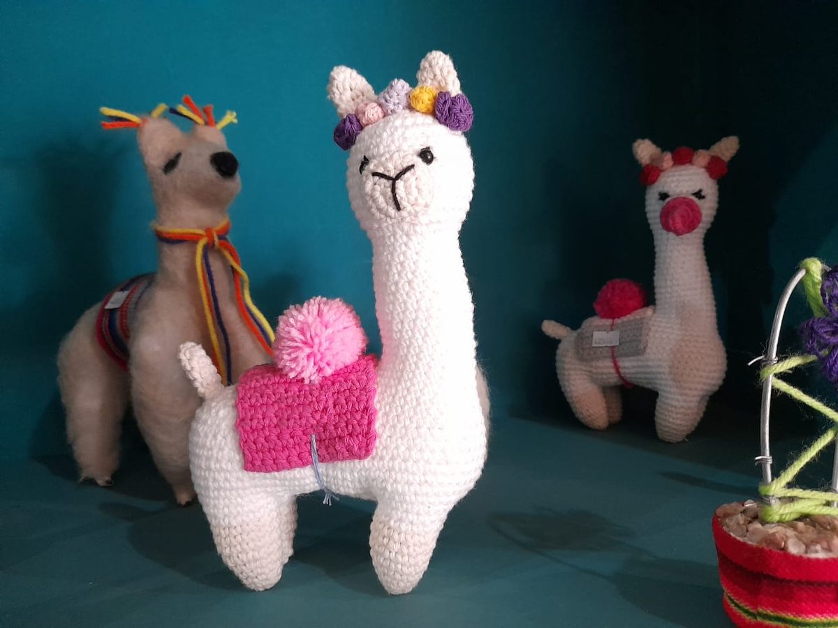 Tres llamitas tejidas al crochet, una blanca con manta rosa y pompón, sobre un estante turquesa